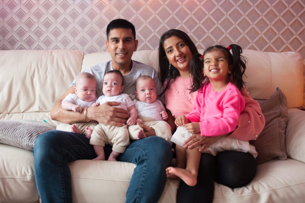 A family (one man, a woman, a child, and baby triplets) sitting on a sofa smiling